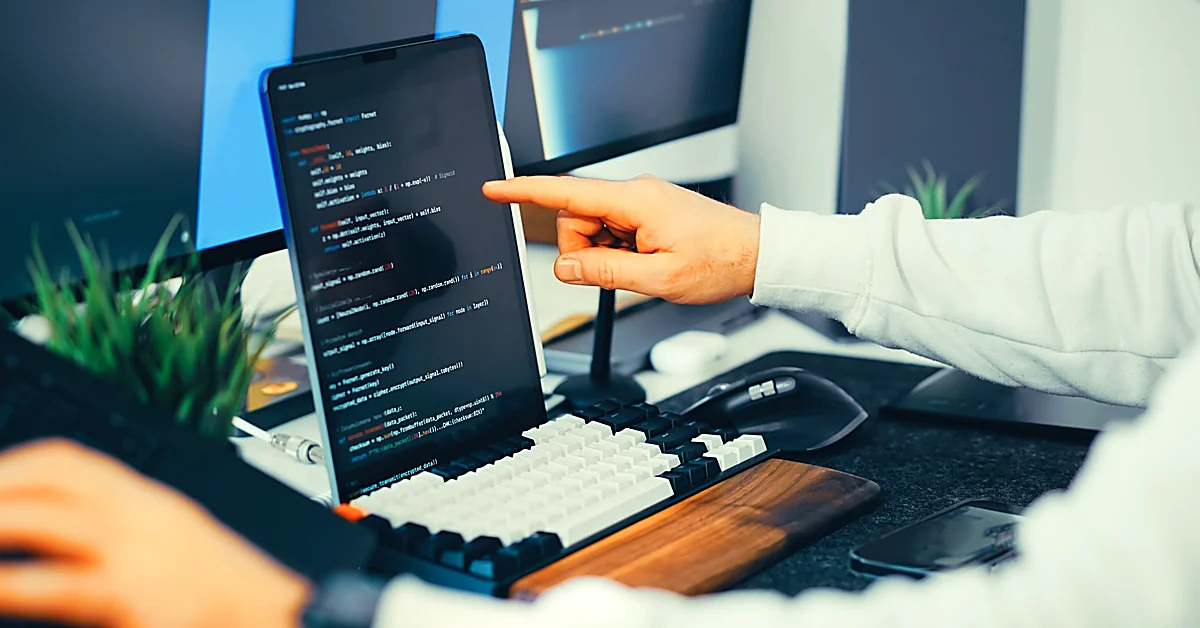 Hand pointing at programming code on a vertical monitor in a developer's workspace with a keyboard and computer equipment.