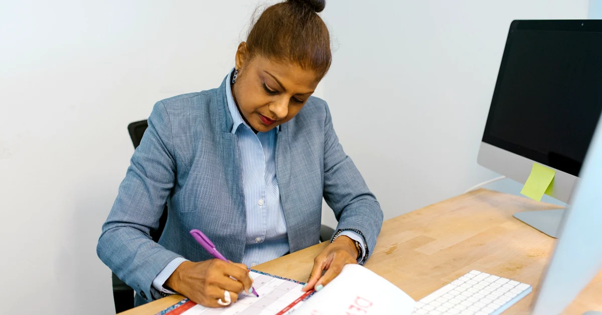Businesswoman in a blue blazer marking a desk calendar with a pen beside a computer in an office.