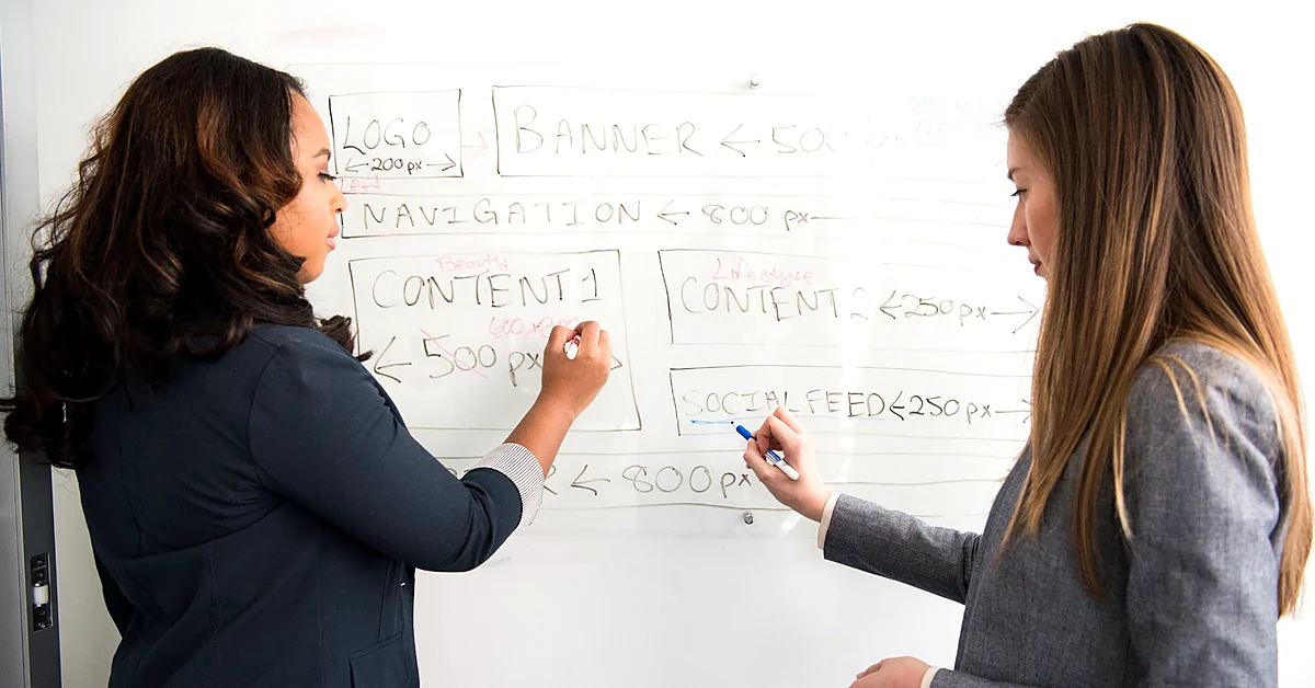 Two women reviewing website navigation and content sections on a whiteboard while planning blog organization.