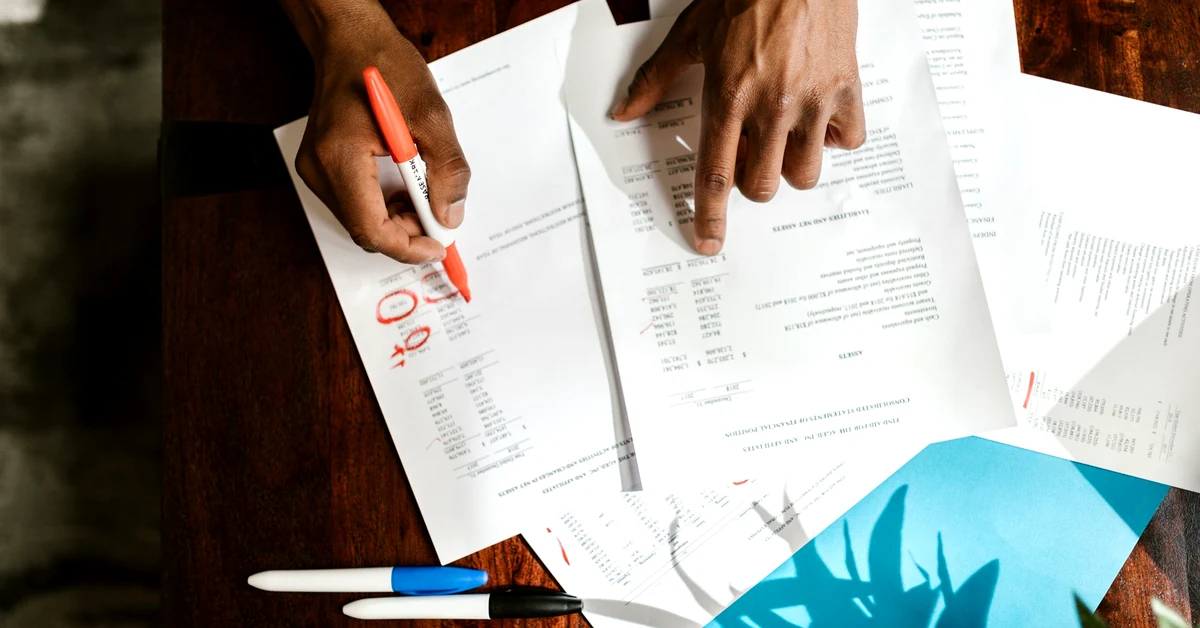 Hands reviewing and marking printed documents during a research and content verification process.
