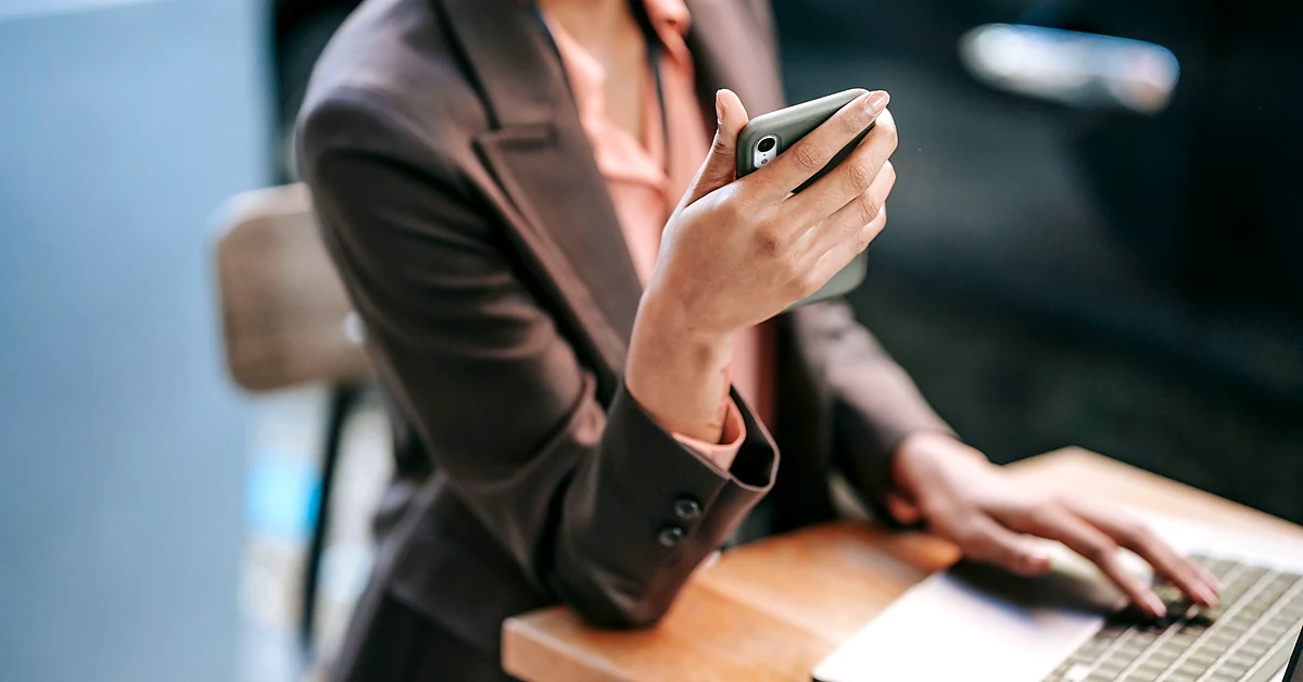 Professional checking a smartphone while working at a laptop, illustrating email signup confirmation testing.