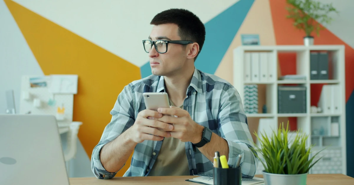 Man checking his smartphone beside an open laptop at a desk