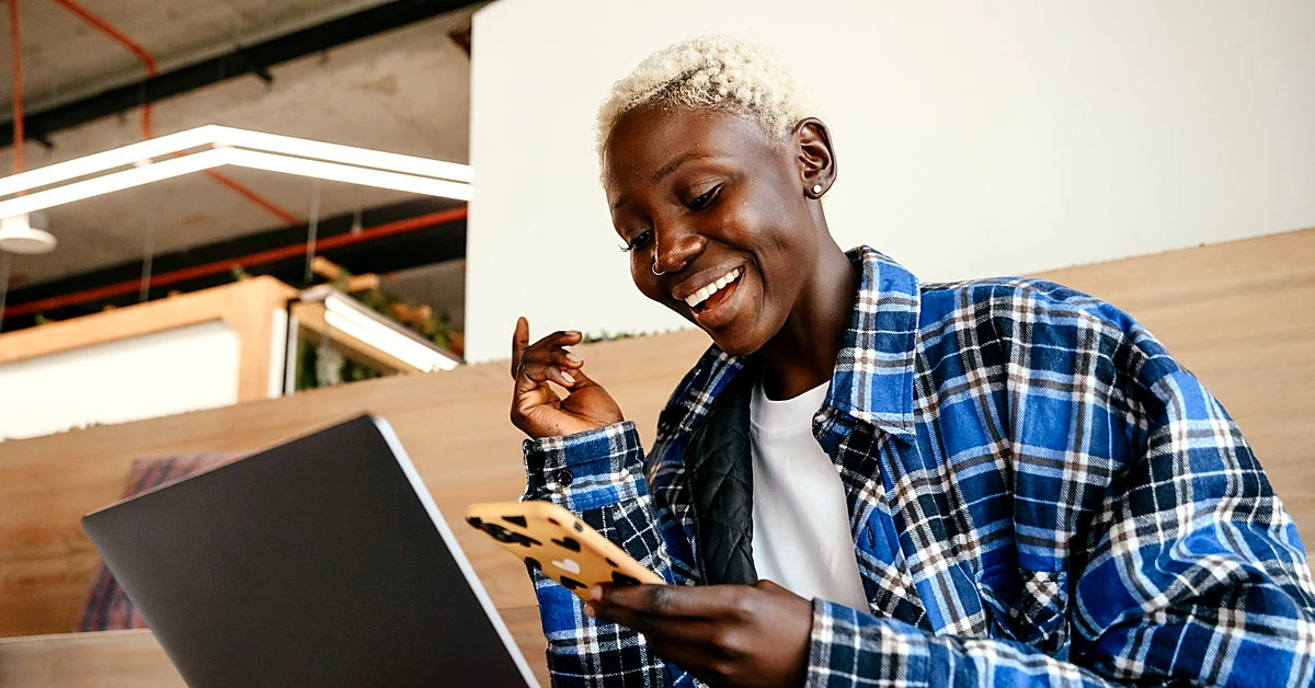 Smiling woman checking her smartphone while working beside a laptop in a modern workspace.