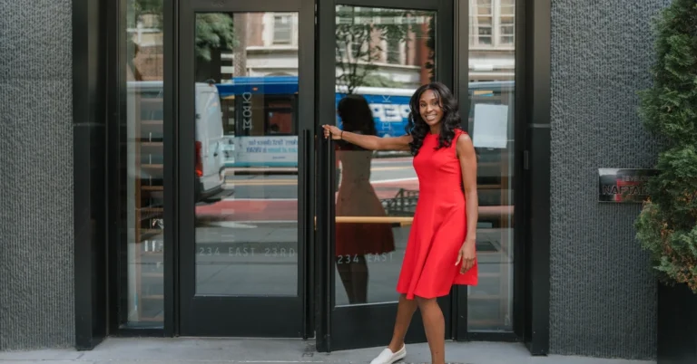 Woman in a red dress holding open a modern office door, illustrating an optional invitation that does not block the visitor’s next step.