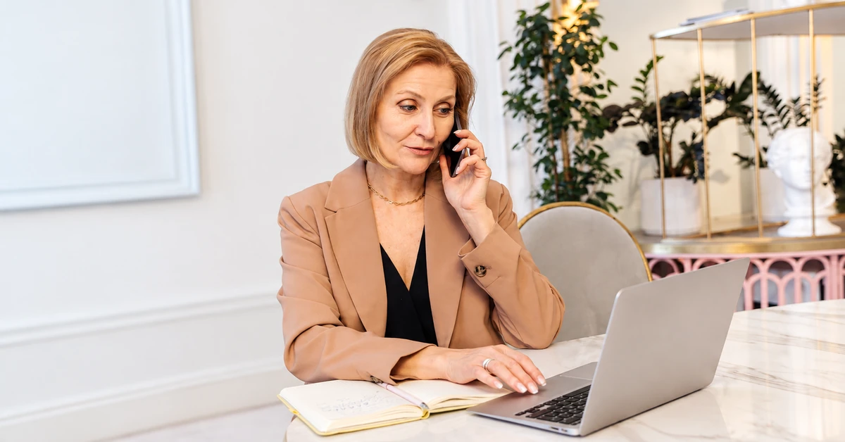 Professional woman in a tan blazer using a laptop while talking on the phone at a desk with a notebook.