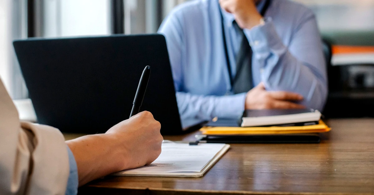 Person completing application paperwork with a pen while another professional reviews information during an office meeting.