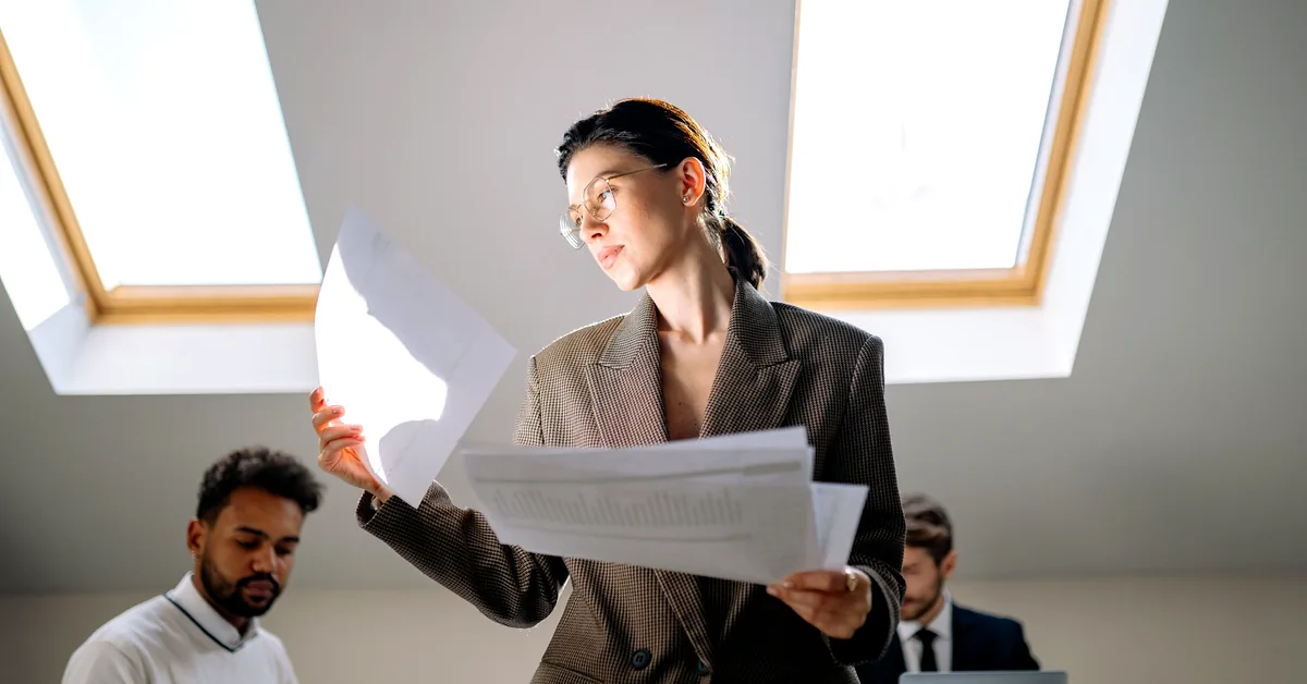 Professional woman wearing glasses reviewing printed documents in a bright office while two colleagues work nearby.