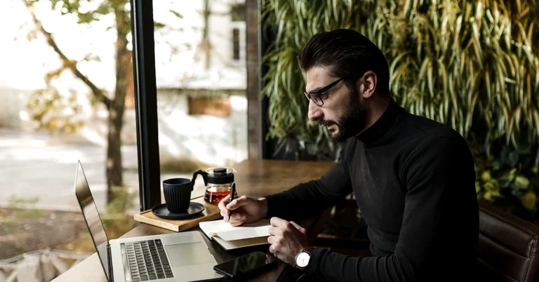 Professional man using a laptop and writing in a notebook while organizing affiliate account setup information at a café.