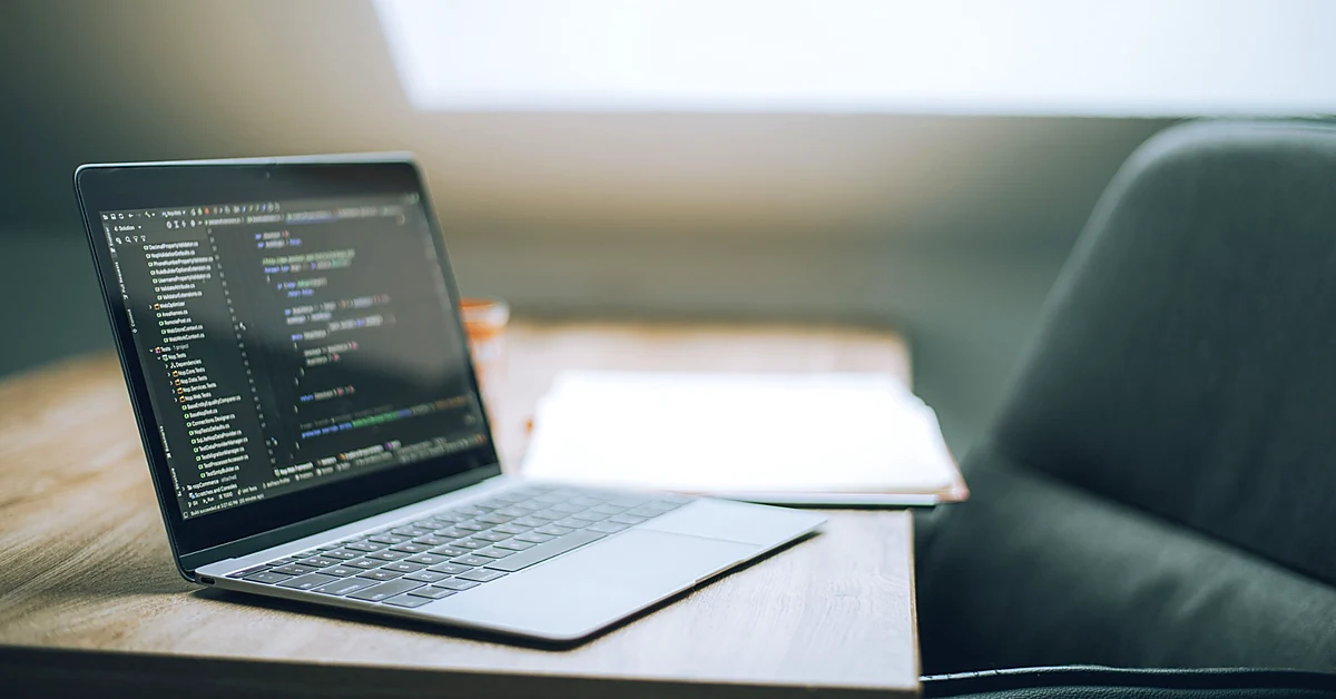 Open laptop displaying programming code on a wooden table beside a notebook in a softly lit workspace.