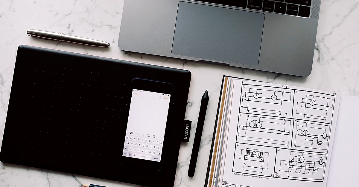 Overhead view of a laptop, drawing tablet, smartphone, and open sketchbook with technical diagrams on a marble desk.
