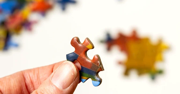 Hand holding a colorful jigsaw puzzle piece with scattered puzzle pieces blurred in the background.