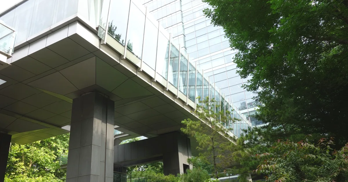 Glass-enclosed pedestrian walkway connecting modern buildings above landscaped greenery.