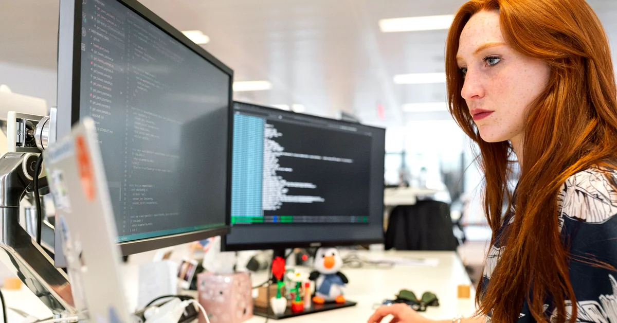 Red-haired woman examining computer code on two monitors at a modern office workstation.