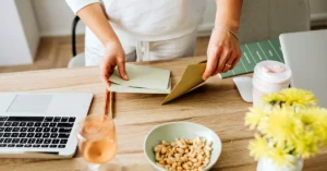 Hands organizing paper envelopes beside a laptop on a wooden desk with flowers and office supplies.