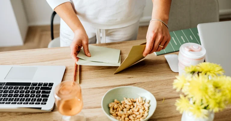 Hands organizing paper envelopes beside a laptop on a wooden desk with flowers and office supplies.