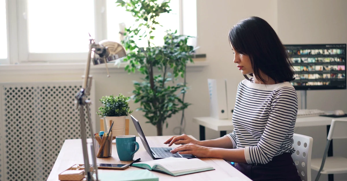 Woman in a black-and-white striped shirt working on a laptop at a bright office desk with a notebook, teal mug, and plants.