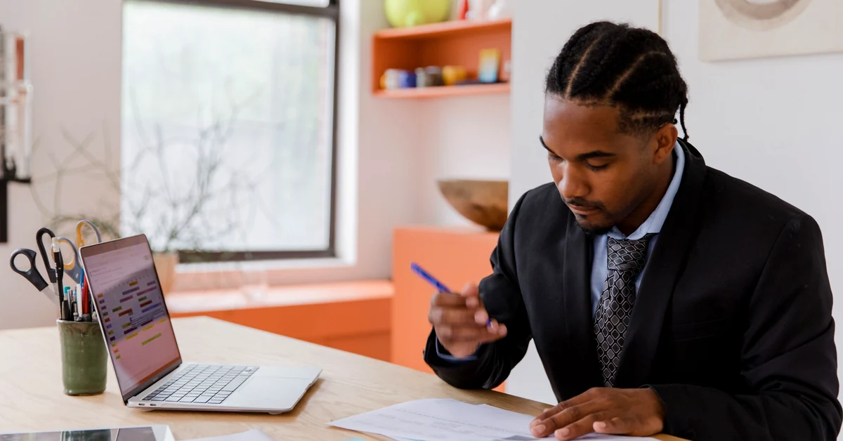 Business professional in a dark suit reviewing printed documents with a pen beside a laptop in a bright office.
