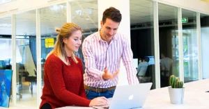 Woman in a rust-colored sweater and man in a pink-striped shirt reviewing a laptop together in a bright modern office.