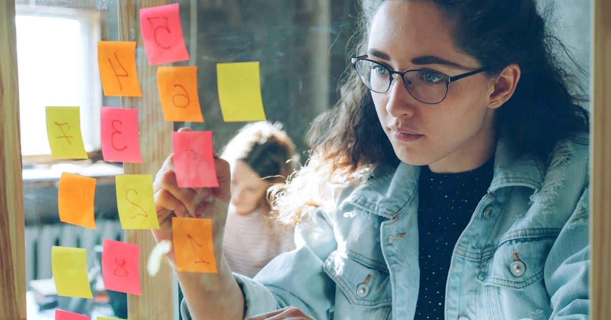Woman wearing glasses arranging colorful sticky notes on a glass wall while planning email strategy steps in an office.