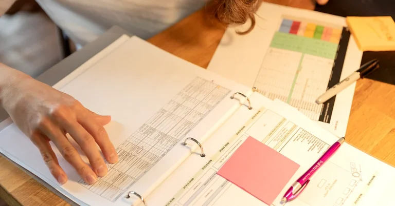 Person reviewing printed documents and sticky notes in an open binder on a wooden desk.