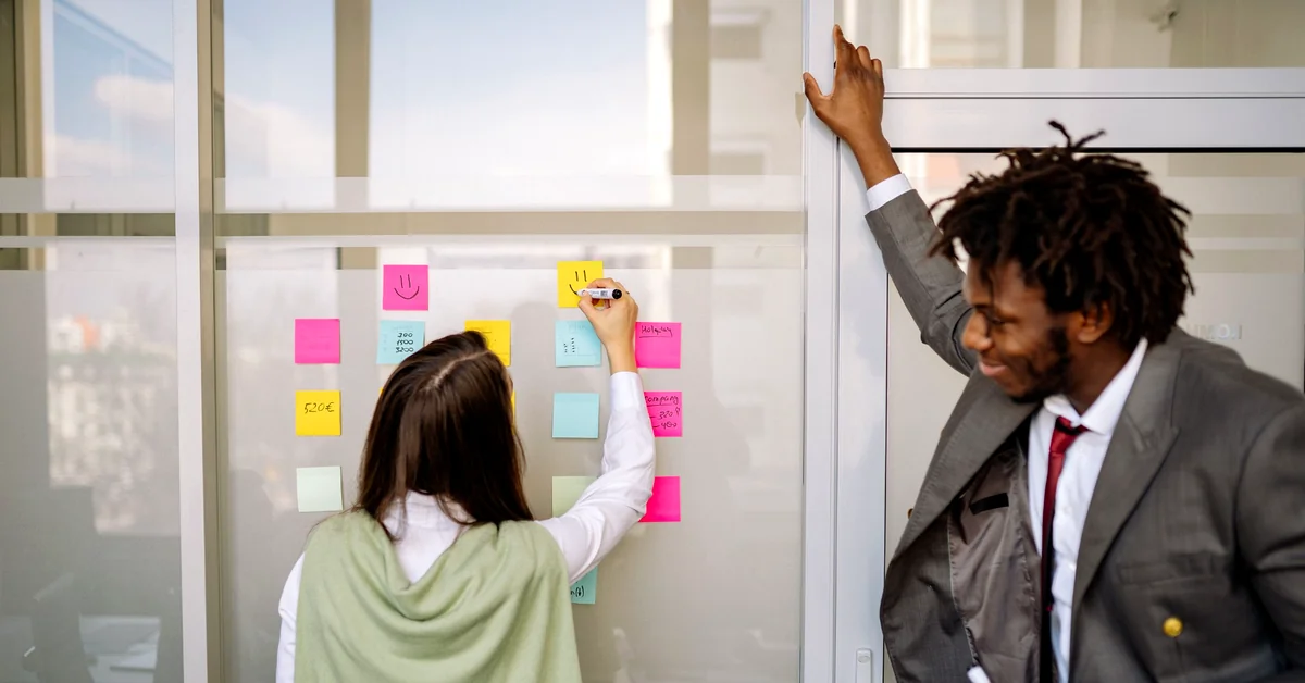 Two colleagues reviewing colorful sticky notes on a glass wall during a collaborative content-planning session.