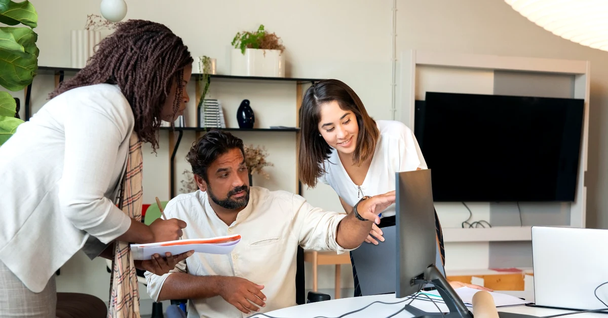 Three colleagues discussing a project around a desktop monitor in a bright office, with one colleague taking notes.