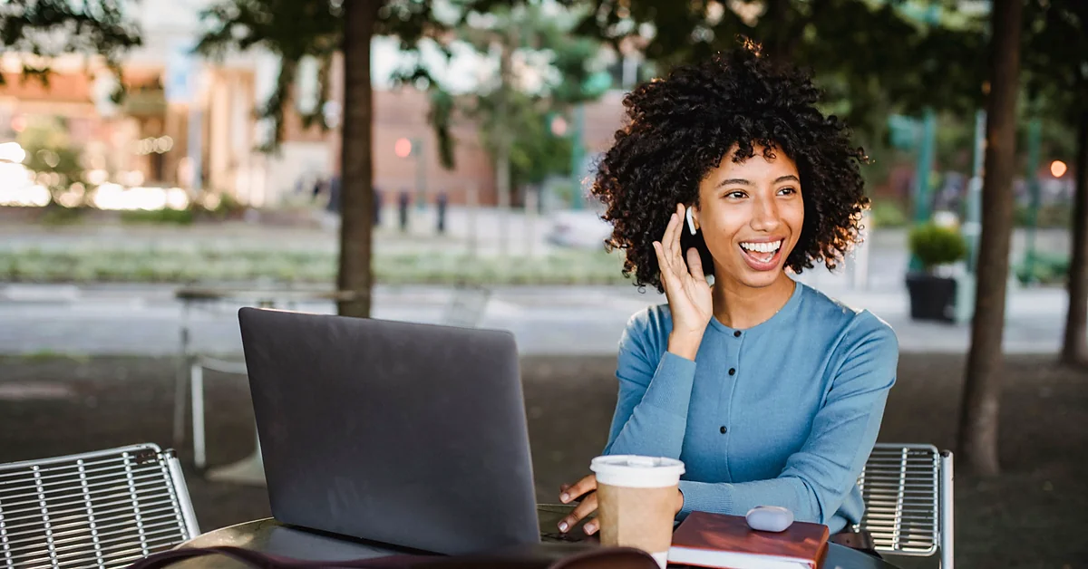 Woman smiling with a wireless earbud beside an open laptop, takeaway coffee, and notebook at an outdoor café.
