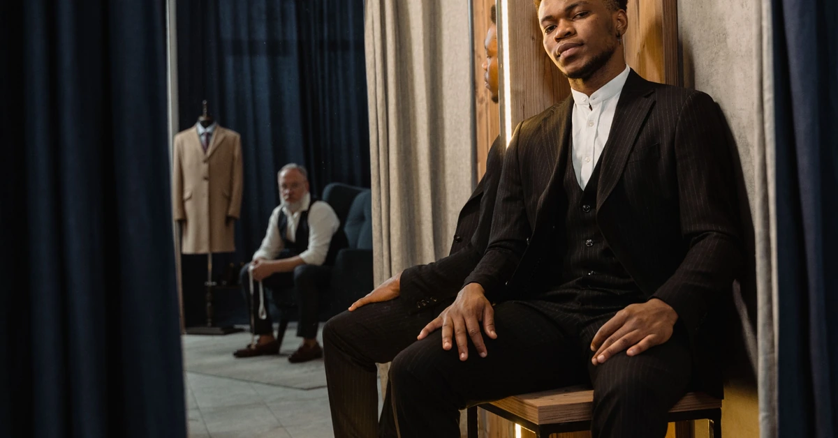 Man seated in a bespoke tailor shop, illustrating how a recognizable personal identity can exist alongside a distinct business brand.