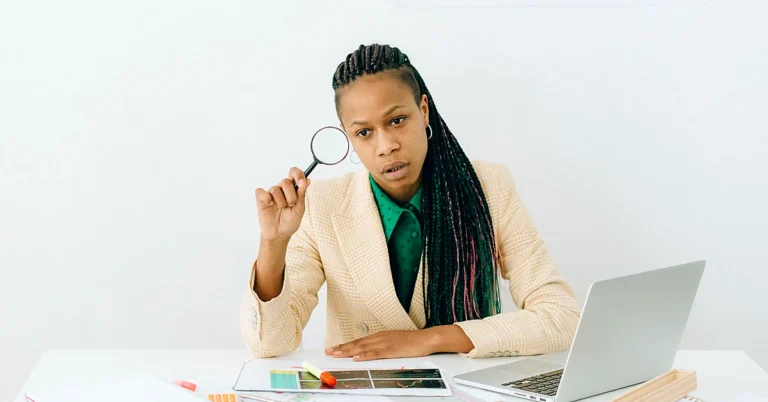 Woman in a cream blazer holding a magnifying glass while examining printed charts beside a laptop.