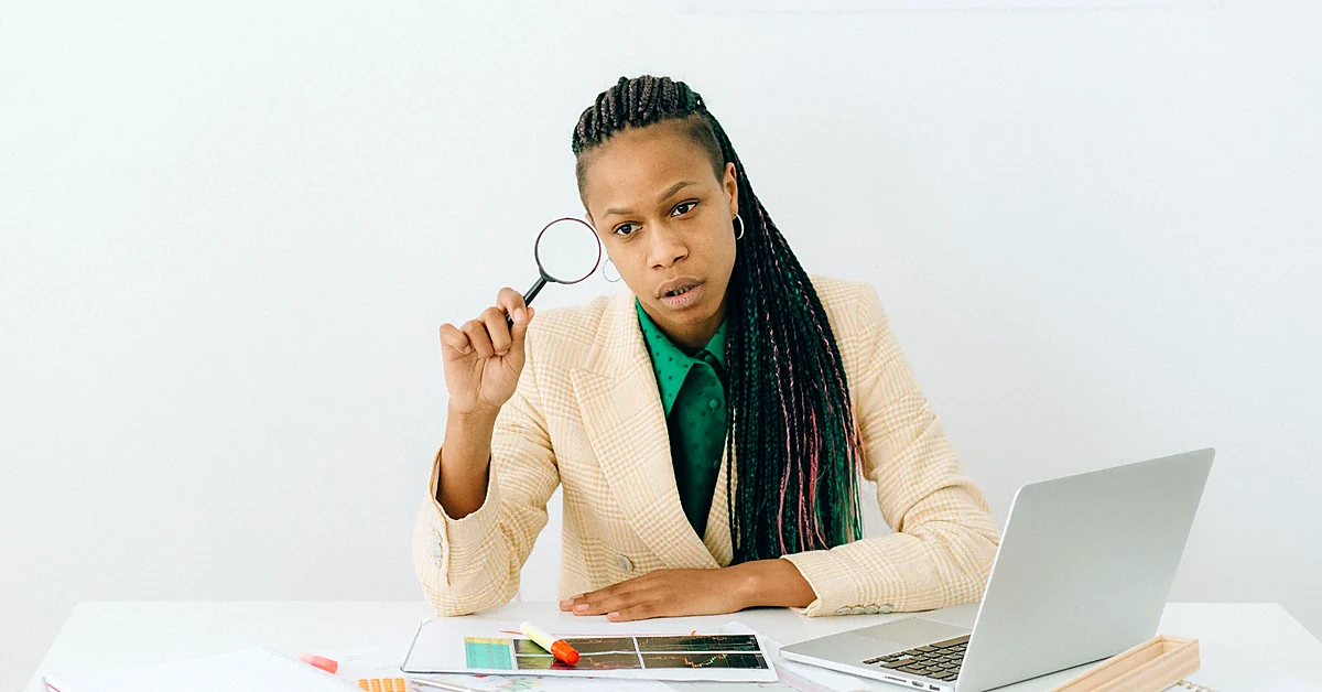 Woman in a cream blazer holding a magnifying glass while examining printed charts beside a laptop.