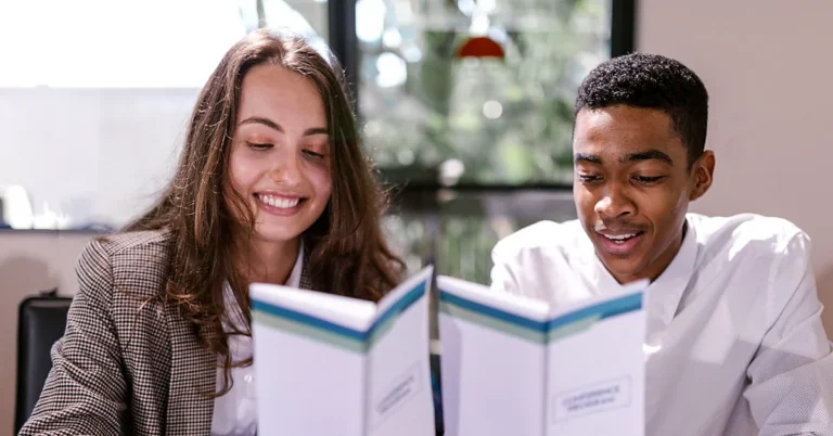 wo colleagues smiling as they review printed booklets together at a table in a bright office.