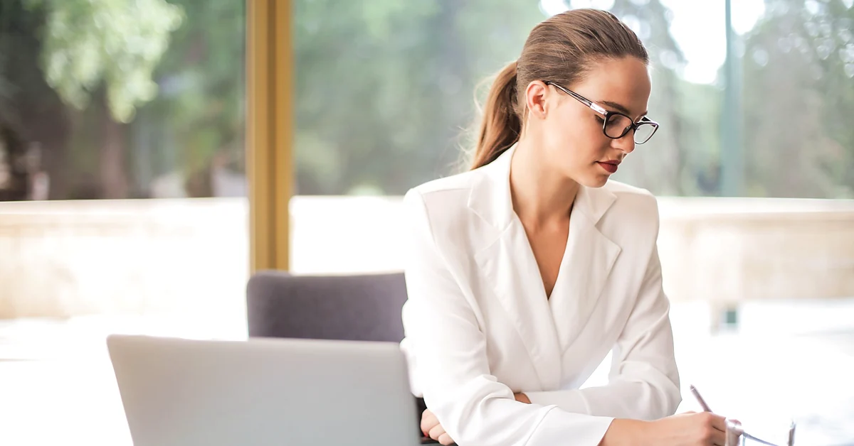 Woman wearing glasses and a white blazer writing at a desk beside an open laptop in a bright office.