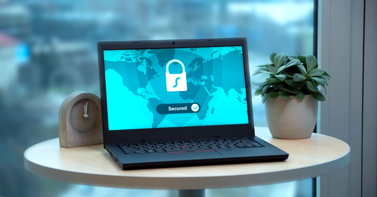Black laptop displaying a security padlock graphic on a round table beside a small clock and potted plant.