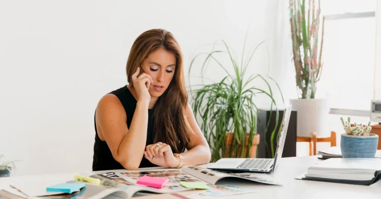 Woman checking her wristwatch while speaking on the phone beside an open laptop at a bright office desk.