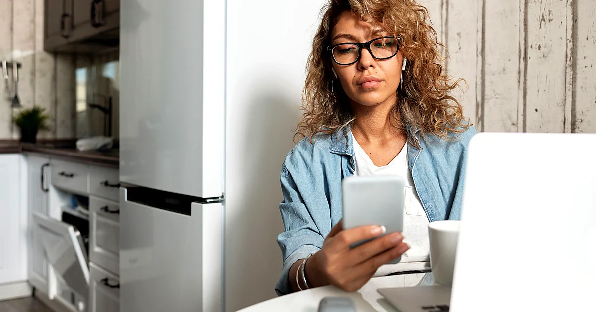 Woman wearing glasses checking a smartphone beside a laptop while reviewing website functionality across devices.