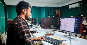 Developer in a plaid shirt working at a computer with programming code and a document displayed on two monitors in a shared office.