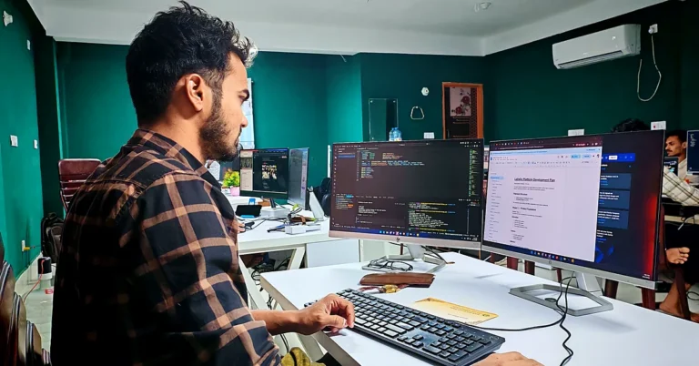Developer in a plaid shirt working at a computer with programming code and a document displayed on two monitors in a shared office.