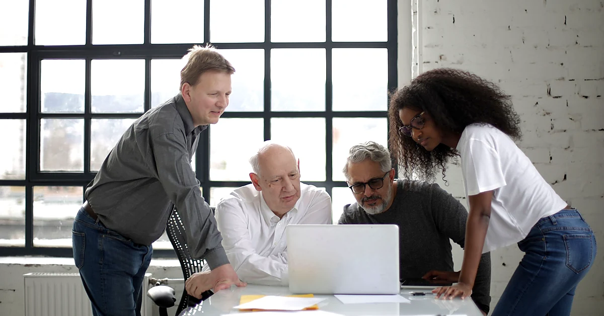 Four colleagues reviewing a project together on a laptop at a bright office conference table.