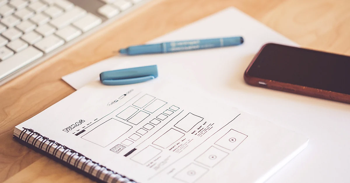 Hand-drawn website wireframe sketches in a notebook beside a smartphone, blue pen, and computer keyboard on a wooden desk.